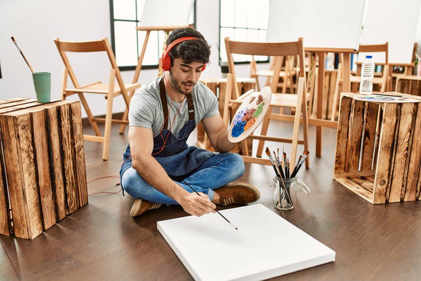 Young hispanic artist man smiling happy and listening to music painting at art studio. - Fotó, kép