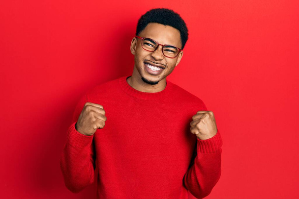 Junger afrikanisch-amerikanischer Mann in lässiger Kleidung und Brille mit erhobenen Armen und geschlossenen Augen, der den Sieg lächelnd feiert. Siegerkonzept.  - Foto, Bild