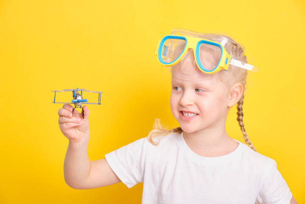 cute caucasian girl toddler with blond hair and pigtails diving mask and a toy airplane on yellow background - Fotografie, Obrázek