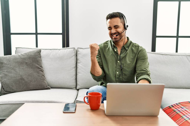Young hispanic man with beard wearing call center agent headset working from home very happy and excited doing winner gesture with arms raised, smiling and screaming for success. celebration concept.  - Zdjęcie, obraz