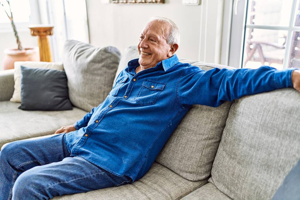 Старший мужчина с седыми волосами сидит на диване в гостиной своего дома. Зрелый мужчина улыбается счастливо дома. - Фото, изображение