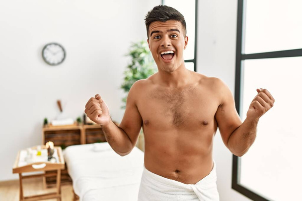 Young hispanic man standing shirtless at spa center celebrating surprised and amazed for success with arms raised and open eyes. winner concept.  - Photo, image