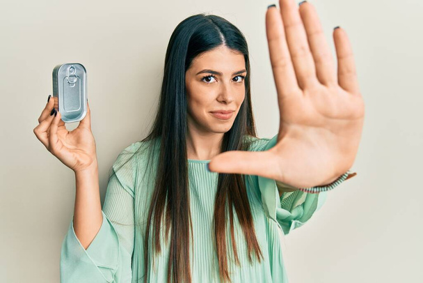 Młoda Latynoska kobieta trzyma konserwy z otwartą ręką robi znak stop z poważnym i pewnym siebie wyrazem twarzy, gest obrony  - Zdjęcie, obraz