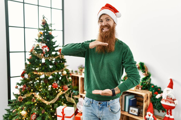 Redhead man with long beard wearing christmas hat by christmas tree gesturing with hands showing big and large size sign, measure symbol. smiling looking at the camera. measuring concept.  - Photo, image