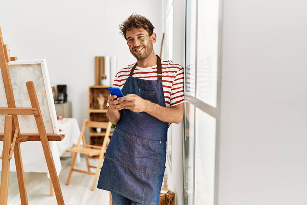 Giovane uomo ispanico sorridente fiducioso utilizzando smartphone in studio d'arte - Foto, immagini