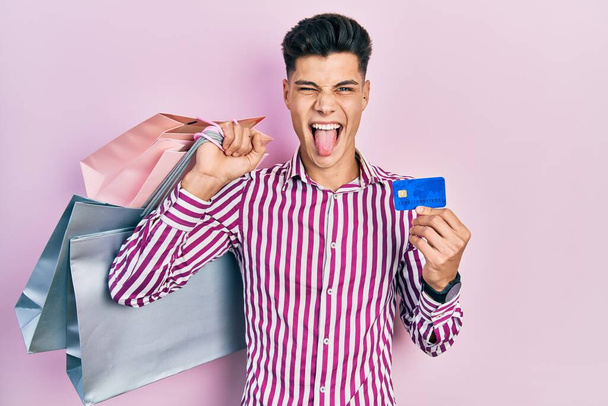 Joven hispano sosteniendo bolsas de compras y tarjeta de crédito sacando la lengua feliz con expresión divertida.  - Foto, imagen