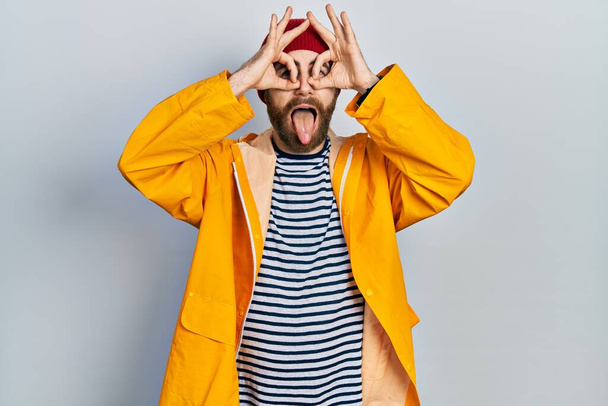 Caucasian man with beard wearing yellow raincoat doing ok gesture like binoculars sticking tongue out, eyes looking through fingers. crazy expression.  - Foto, Imagen