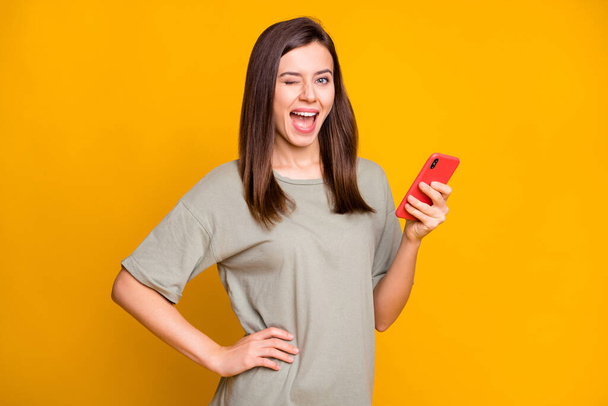 Photo of young attractive pretty happy smiling excited girl wink blink eye hold phone isolated on yellow color background. - Foto, Imagem