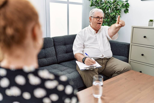 Senior psychologist man at consultation office pointing with finger surprised ahead, open mouth amazed expression, something on the front  - Photo, Image