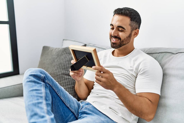 Jeune homme hispanique souriant confiant regarder cadre photo à la maison - Photo, image
