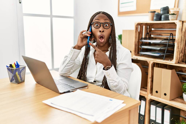 Czarna kobieta z warkoczami pracująca w biurze, mówiąca przez telefon, zaskoczona wskazaniem palcem w bok, otwarte usta, zdumiona ekspresja.  - Zdjęcie, obraz