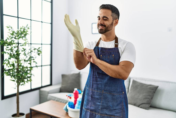 Joven hombre hispano sonriendo confiado usando delantal y limpiando guantes en casa - Foto, Imagen