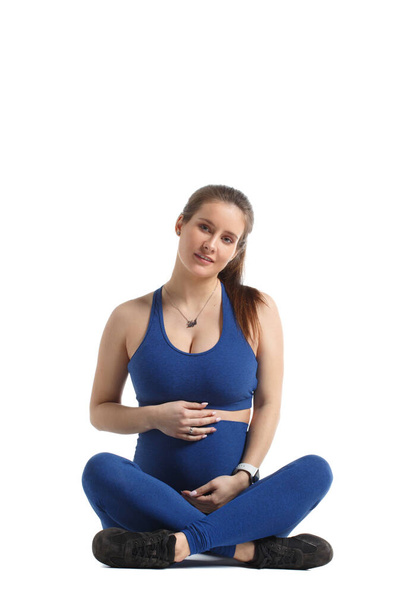 Söpö raskaana oleva nainen sinisissä urheiluvaatteissa istuu eristyksissä valkoisella taustalla. - Valokuva, kuva