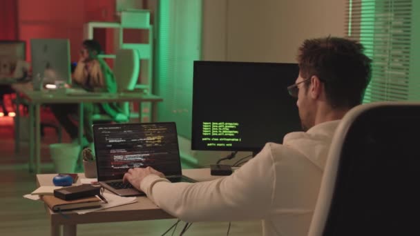 Vista trasera apuñalada de código de programa de escritura hacker tanto en el ordenador portátil y PC sentado en el escritorio en la oficina con iluminación roja y verde junto con colega afroamericano - Imágenes, Vídeo