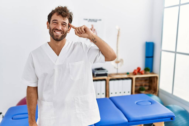 Young handsome physiotherapist man working at pain recovery clinic smiling pointing to head with one finger, great idea or thought, good memory  - Foto, Imagem