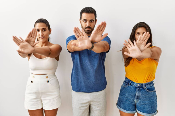 Grupo de jóvenes hispanos de pie frente a aislada expresión de rechazo cruzando brazos y palmas haciendo signo negativo, cara enojada  - Foto, imagen