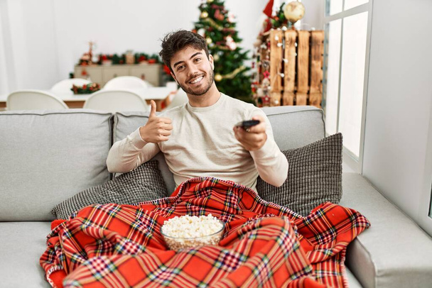 Νεαρός Ισπανός που τρώει ποπ κορν καθισμένος στον καναπέ δίπλα στο χριστουγεννιάτικο δέντρο χαμογελώντας χαρούμενος και θετικός, με τον αντίχειρα να κάνει εξαιρετική δουλειά και το σήμα έγκρισης  - Φωτογραφία, εικόνα