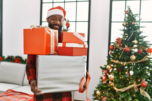 Giovane uomo afroamericano in possesso di regali in piedi vicino all'albero di Natale a casa - Foto, immagini