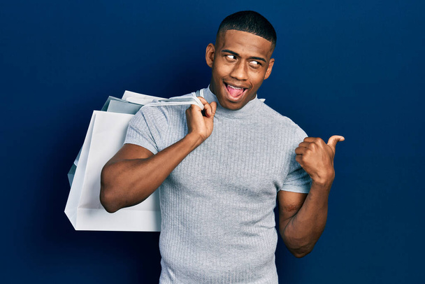Young black man holding shopping bags pointing thumb up to the side smiling happy with open mouth  - 写真・画像