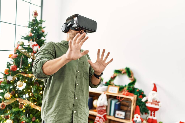 Young hispanic man using vr goggles standing by christmas tree at home - Photo, Image