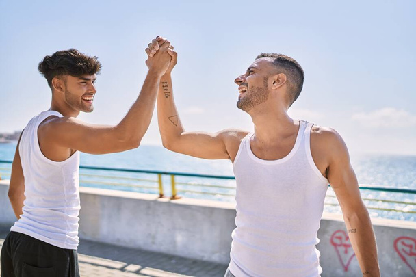 Два латиноамериканских мужчины улыбаются уверенно пять с поднятыми руками на берегу моря - Фото, изображение