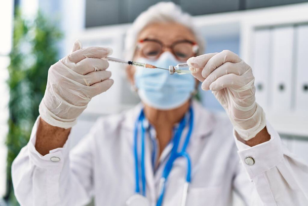 Mujer mayor de cabello gris que usa uniforme médico y máscara médica con dosis de vacuna en la clínica - Foto, Imagen