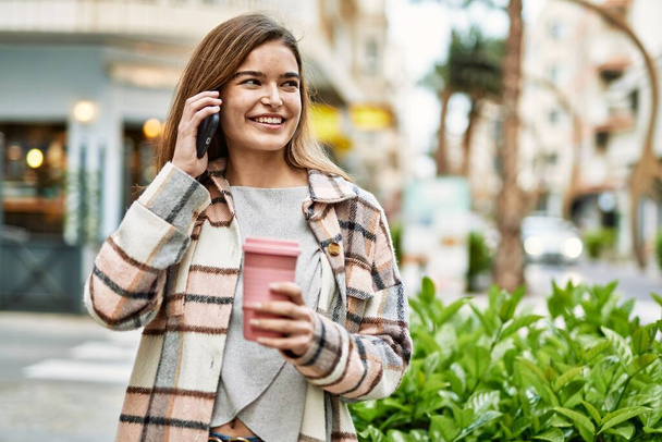 Young blonde woman holding coffee talking on the smartphone at street - Foto, imagen