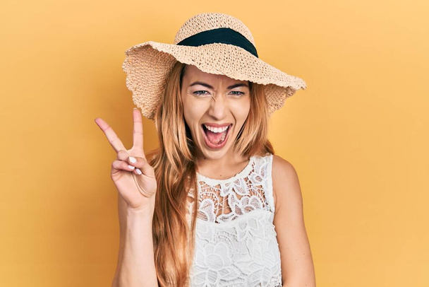 Jeune femme caucasienne portant le chapeau d'été souriant avec le visage heureux clin d'oeil à la caméra faisant signe de victoire. numéro deux.  - Photo, image