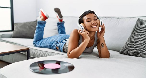 Mujer hispana joven escuchando música tumbada en un sofá en casa - Foto, imagen