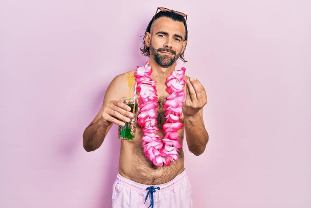 Young hispanic man wearing swimsuit and hawaiian lei drinking tropical cocktail doing money gesture with hands, asking for salary payment, millionaire business  - Photo, Image