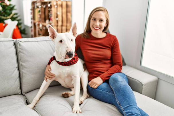 Young caucasian woman smiling confident hugging dog sitting by christmas tree at home - Photo, image