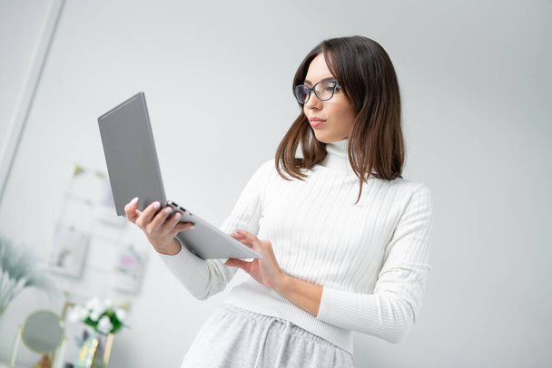 Business woman in glasses stands home and looks into laptop screen. - Zdjęcie, obraz