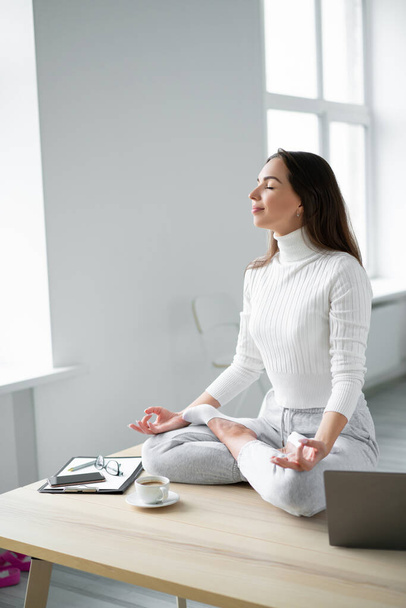 Young woman sitting on desk  in padmasana yoga pose home office. - Zdjęcie, obraz