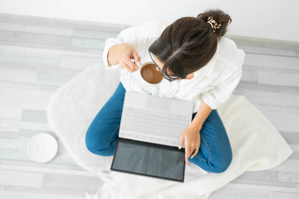Woman in white shirt sits on floor home with laptop and drinks coffee. - Photo, image
