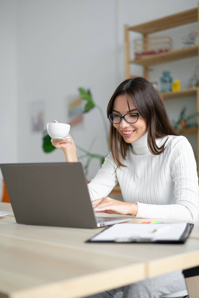 Close up of woman holding coffee looking laptop in Front of her in white room. - Photo, Image