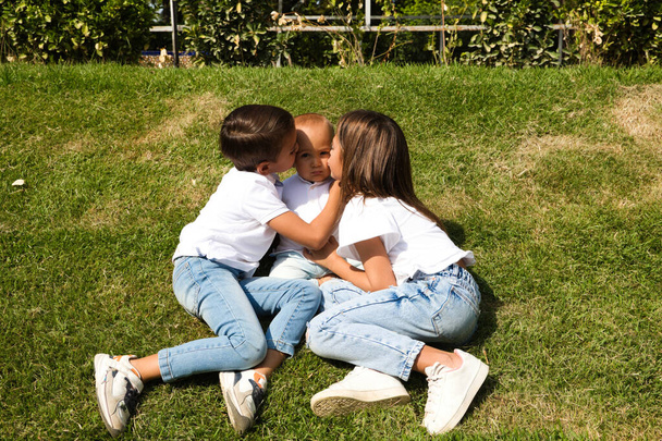 Retrato de tres hermanos pequeños besando a su hermano pequeño en el parque. Están vestidos con camiseta blanca y jeans. Concepto de una familia unida y feliz. - Foto, imagen