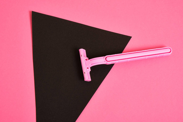 a pink plastic disposable razor and a black triangle made of paper on a pink background, an association on the theme of feminism and shaving hair from a female body - Foto, Bild