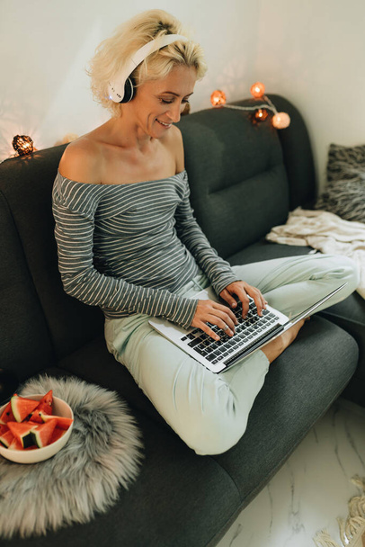 Adult blonde woman in headphones sitting on a sofa and working by laptop from home - Foto, Imagem