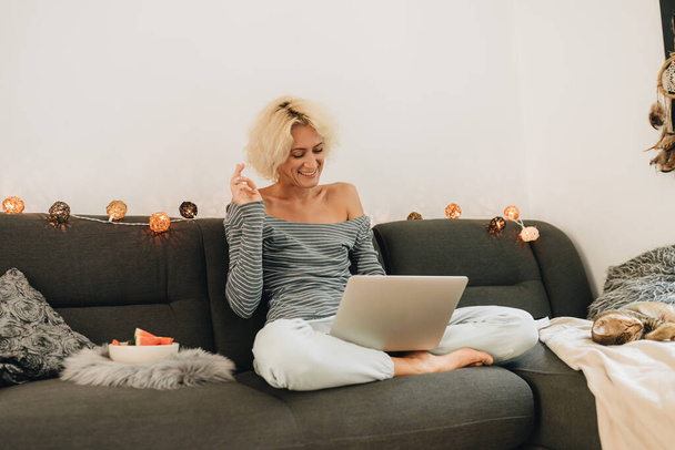 Adult blonde woman in headphones sitting on a sofa and having video chat by laptop from home - Foto, Imagem