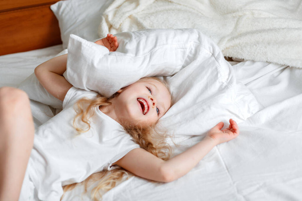Cheery little Kid girl having fun jumping in living room playing with pillow. Portrait of preschool daughter Caucasian blonde girl in pajamas play in bed - Photo, image