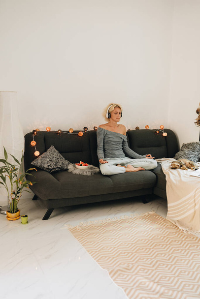 Felnőtt nő fülhallgatóban ül egy kanapén otthon és meditál - Fotó, kép