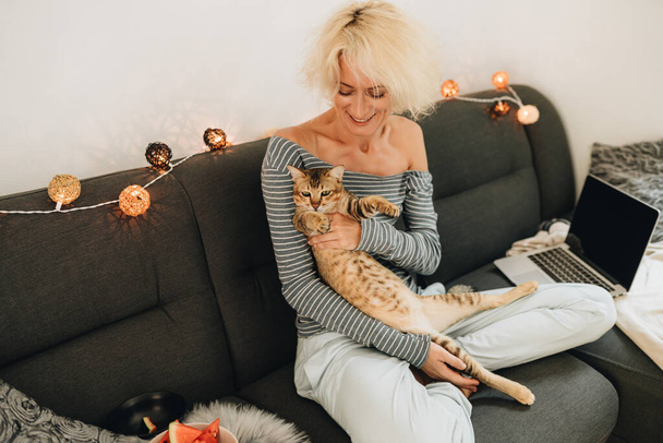 Mujer rubia adulta sentada en un sofá y jugando con un gato - Foto, imagen