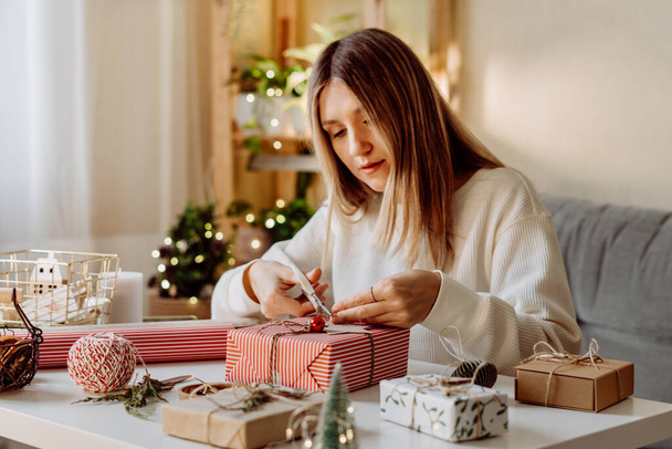 Nő csomagolás karácsonyi öko-természetes ajándék dobozok otthon. Ajándékok elkészítése fehér asztalon díszítő elemekkel, fényekkel és tárgyakkal Karácsonyi vagy újévi barkácsolás koncepció - Fotó, kép