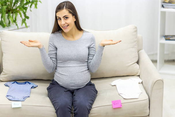Photo of pregnant woman sitting on the sofa with child clothes. - Foto, afbeelding