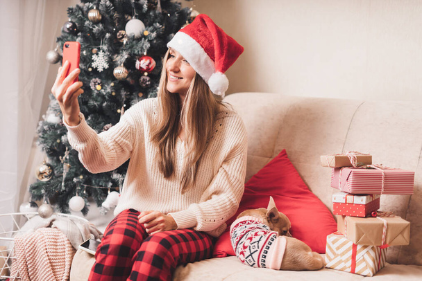 Fiatal nő és kutya pulóverben szelfi portrét készít az okostelefonon a téli szünetben, élvezze a karácsonyi időt otthon. Csomagolás ajándékok és video chat a mobiltelefon - Fotó, kép