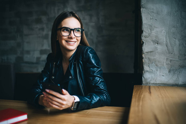Happy Caucaisan hipster girl in optical spectacles for provide eyes correction enjoying leisure pastime in coffee shop, cheerful female blogger with modern cellphone device smiling indoors - Foto, afbeelding