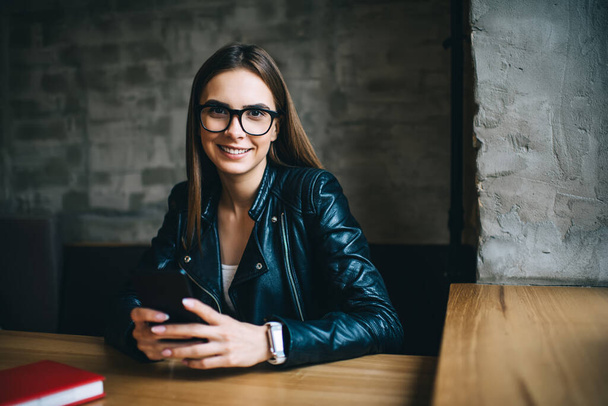Happy Caucaisan hipster girl in optical spectacles for provide eyes correction smiling at camera during leisure time in coffee shop, portrait of cheerful female blogger with modern cellphone device - Zdjęcie, obraz