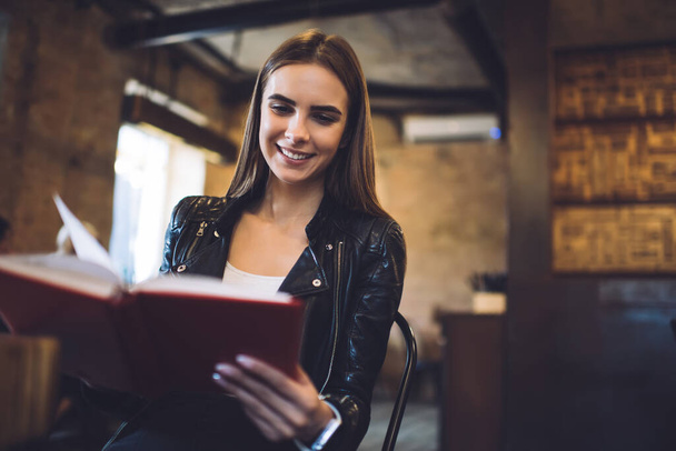 Happy Caucasian kobieta czyta pozytywne powieści artykuł uśmiechając się i ciesząc rozrywki w kawiarni wnętrza, beztroski hipster dziewczyna z literatury bestseller uczucie beztroski z ulubionym hobby - Zdjęcie, obraz