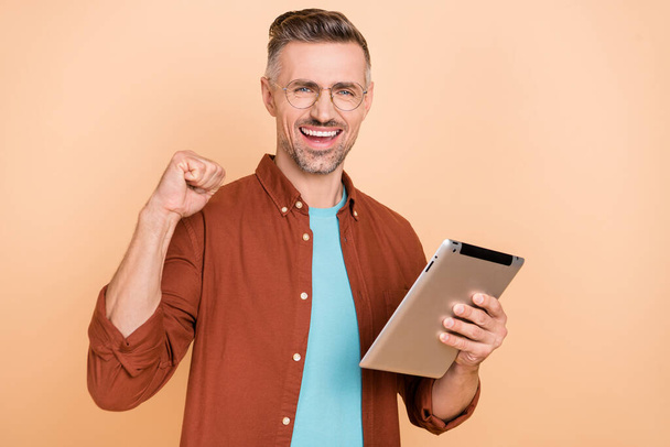 Photo of delighted middle-aged guy use gadget application raise fists up shopping sale isolated over pastel beige color background. - Фото, зображення