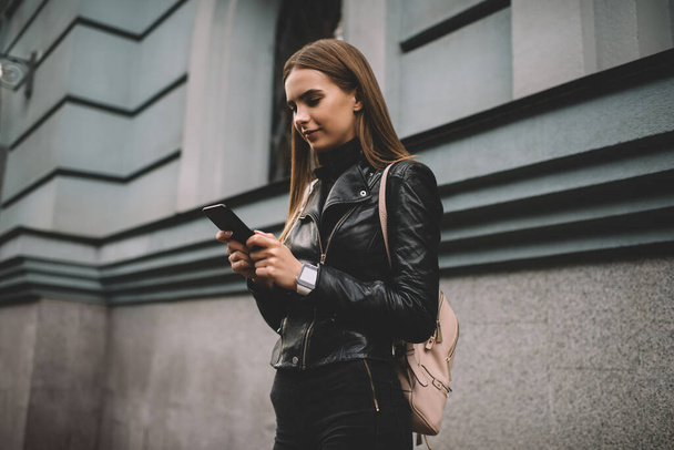 Caucasian tourist tracking GPS via location application on modern cellphone device, trendy dressed hipster girl in leather jacket reading travel publication while browsing social media on smartphone - Foto, Imagen